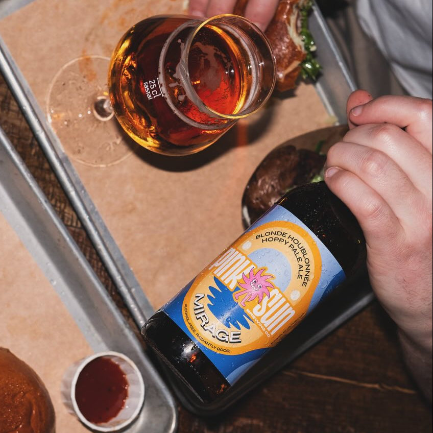 Person holding a bottle of beer with a colorful label, surrounded by food on a tray.